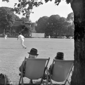 Prints of KEW GREEN, London. Rear view of an elderly couple seated on deckchairs watching a cricket match being played on Kew Green. John Gay. Date range: Jan 1962 - May 1964 Sport Prints: Print of an elderly couple watching a cricket match on Kew Green