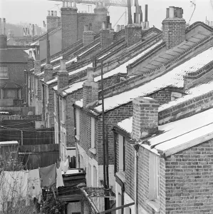 Prints of A view from roof level along the snowy roofs of a line of terraced brick houses, showing chimneys, the back gardens of the houses, and with a large crane visible in the distance above the roofs House And Home Prints: Rooftops Print: A snowy view of brick houses and a distant crane
