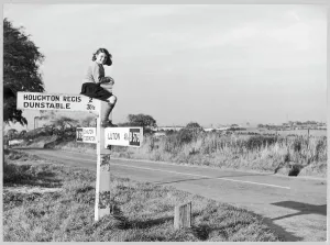 Prints of A girl in sturdy shoes and long socks, probably school uniform, perches on top of a signpost by the B579 road near Chalton in Bedfordshire Bedfordshire Prints: Bedfordshire Road Signs Print 1950s Childhood Scene