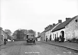 Prints of Bellaghy, Co. Derry - a street scene with motor car in the middle of the road and people to the left. There are thatched buildings. (Location: Northern Ireland: County Londonderry: Bellaghy) County Londonderry Prints: Early 20th Century Bellaghy Street Scene Print
