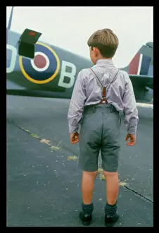 Prints of Spitfire Summer: A little boy wearing shorts and braces admires a Spitfire on the runway at Biggin Hill, Kent, England Aviation Prints: Spitfire Print: Boy Admires a Spitfire on the Runway