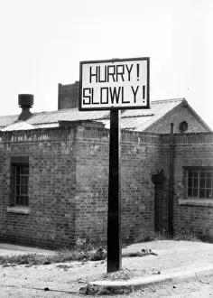Prints of Hurry! Slowly! This curiously worded signpost is to be seen by the roadside near Ashby-de-la-Zouche, Leicestershire, England. It warns of a open Level Crossing Date: 1950s Leicestershire Prints: 1950s Leicestershire Level Crossing Print: HURRY! SLOWLY