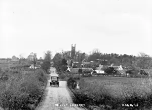 Prints of The Loup, Co. Derry - a view of a village beside a road with a motor car and people in the foreground. There are thatched buildings and a church Derry Prints: Early 20th Century Print of The Loup Village, Northern Ireland