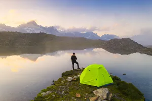 Rear view of man with hat standing outside tent at Obere Schwarziseeli lake at dawn Rear view of man with hat standing outside tent at Obere Schwarziseeli lake at dawn