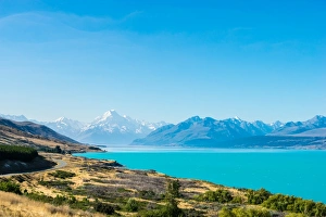 A road winds along the edge of a turquoise blue lake with mountains in the distance A road winds along the edge of a turquoise blue lake with mountains in the distance
