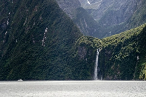 A sightseeing ship dwarfed by a tall waterfall in a fjord, South Island, New Zealand A sightseeing ship dwarfed by a tall waterfall in a fjord, South Island, New Zealand