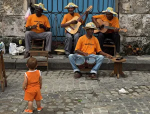 Street band wearing orange shirts playing music on the pavement watched by toddler wearing orange clothes Street band wearing orange shirts playing music on the pavement watched by toddler wearing orange clothes