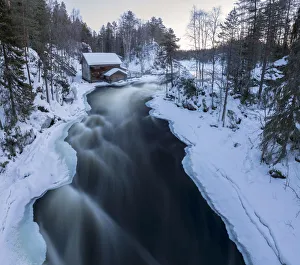 A wooden hut surrounded by the river rapids and snowy woods at dusk Juuma Myllykoski A wooden hut surrounded by the river rapids and snowy woods at dusk Juuma Myllykoski