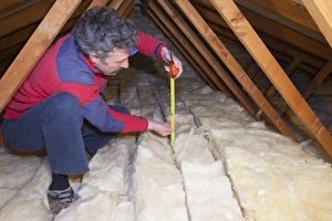 A man measuring the depth of insulation in a house loft or roof space. Insulating your loft can save a significant A man measuring the depth of insulation in a house loft or roof space. Insulating your loft can save a significant
