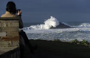 A woman takes pictures of waves as they crash against a lighthouse in the port town of A woman takes pictures of waves as they crash against a lighthouse in the port town of