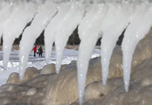 Two women are framed by icicles and mounds of frozen ice as they walk along a beach in Two women are framed by icicles and mounds of frozen ice as they walk along a beach in