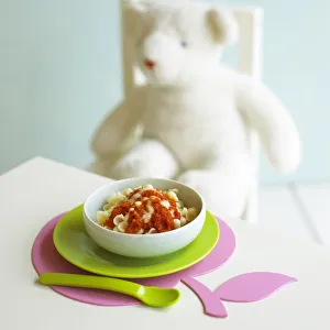 Bowl of pasta with tomato and cheese sauce on table, teddy bear sitting on chair in background Bowl of pasta with tomato and cheese sauce on table, teddy bear sitting on chair in background