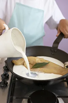 Girl pouring milk into frying pan containing piece of haddock and bay leaf Girl pouring milk into frying pan containing piece of haddock and bay leaf