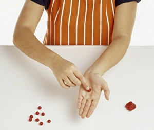 Hand model wearing orange and white striped apron, rolling red marzipan balls in palm of hand
