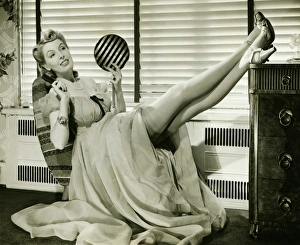 Glamorous woman in evening gown holding hand mirror, legs on cabinet, (B&W), portrait Glamorous woman in evening gown holding hand mirror, legs on cabinet, (B&W), portrait