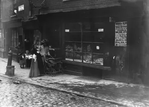 Prints of 1907: Women pushing a pram past a bakery window in Little Italy, a slum area of London in the borough of Holborn. (Photo by Topical Press Agency/Getty Images) Slums Prints: Print of Women and Child in Little Italy Slum, London