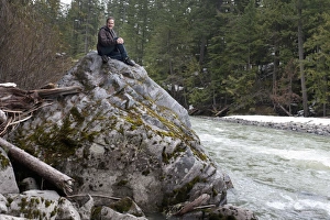 A Man Sits On A Large Rock Elevated Above The River Flowing Below In Jasper National Park A Man Sits On A Large Rock Elevated Above The River Flowing Below In Jasper National Park