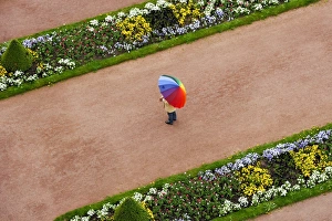 Person with umbrella in rainbow colors on a red gravel path between flower beds in Person with umbrella in rainbow colors on a red gravel path between flower beds in