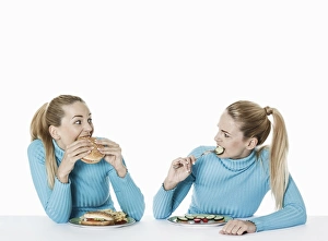 Two women eating food, one eating fast food, the other one eating uncooked vegetarian food Two women eating food, one eating fast food, the other one eating uncooked vegetarian food