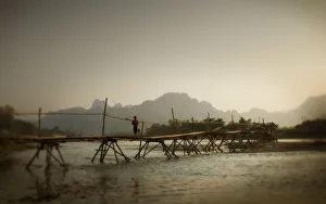 Boy Running Across A Wooden Bridge On A River With Mountains In The Background; Vang Boy Running Across A Wooden Bridge On A River With Mountains In The Background; Vang
