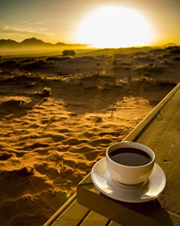 A cup of coffee sits on a wooden table with a view of the sunrise over the desert, Namibia A cup of coffee sits on a wooden table with a view of the sunrise over the desert, Namibia