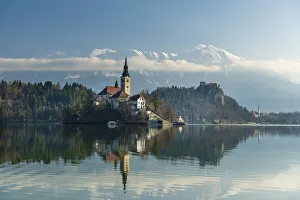 Looking across the calm waters of Lake Bled at dawn to the church on Lake Bled Island and Bled Castle behind; Bled Looking across the calm waters of Lake Bled at dawn to the church on Lake Bled Island and Bled Castle behind; Bled