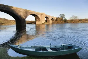 Northumberland, England; A Boat Moored On The Edge Of A River With A Bridge Northumberland, England; A Boat Moored On The Edge Of A River With A Bridge