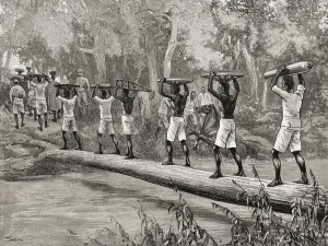 Porters carrying dismantled guns across a stream in the Niger Territories in an expedition against slave raiders Porters carrying dismantled guns across a stream in the Niger Territories in an expedition against slave raiders