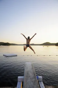 A Swimmer Jumps Off A Diving Board As The Sun Sets Over A Lake In Muskoka; Huntsville, Ontario, Canada A Swimmer Jumps Off A Diving Board As The Sun Sets Over A Lake In Muskoka; Huntsville, Ontario, Canada