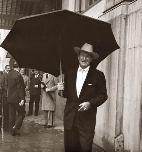 Prints of John Wayne standing under a big black umbrella in the rain in London October 1960 Weather Prints: John Wayne Under Black Umbrella Print London 1960