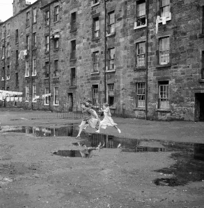 Prints of Three young friends leap over a puddle outside a Govan tenement block in Glasgow. September 1956 United Kingdom Prints: Three Young Friends Leap Over a Puddle Print, Glasgow 1956