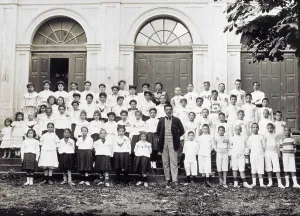 Portrait of an elementary school group. The little girls are wearing school uniforms and the boys are wearing sport uniforms. At the center of the group, a teacher