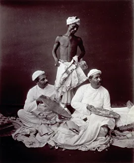 Three traders in Colombo (Ceylon) surrounded by numerous pieces of cloth; two men are sitting and displaying two finely worked trays to another person who is out of view. Behind them, an indian servant
