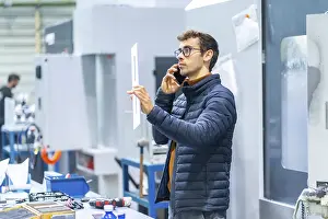 Communicative Young Adult Man Prints: Engineer talking to the mobile in a computer numeric control factory