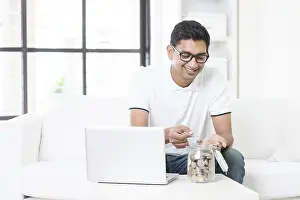 Man Relaxing At Home Prints: Indian guy using internet computer and counting coins at home