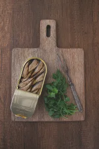 Prints of An open can of sprats, appetizer, on a wooden cutting board, with a fork and herbs, top view Sprat Prints: An Open Can of Sprats Print, Top View