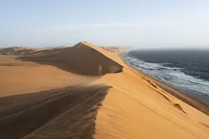 Prints of Sand dunes with wave patterns and sea in evening light, wind blowing sand across the dunes, Atlantic coast and sandy desert with sea of dunes, impressive desert landscape at sunset, Sandwich Harbour Heritage Site Prints: Sand Dunes Print - Namib Desert Evening Scene