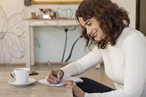 Female Entrepreneurship Prints: Smiling entrepreneur writes notes in a notebook while working at a table