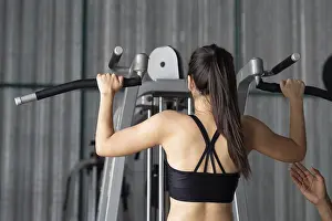 Wellness And Health Prints: A young woman performs pull-ups with the help of her trainer